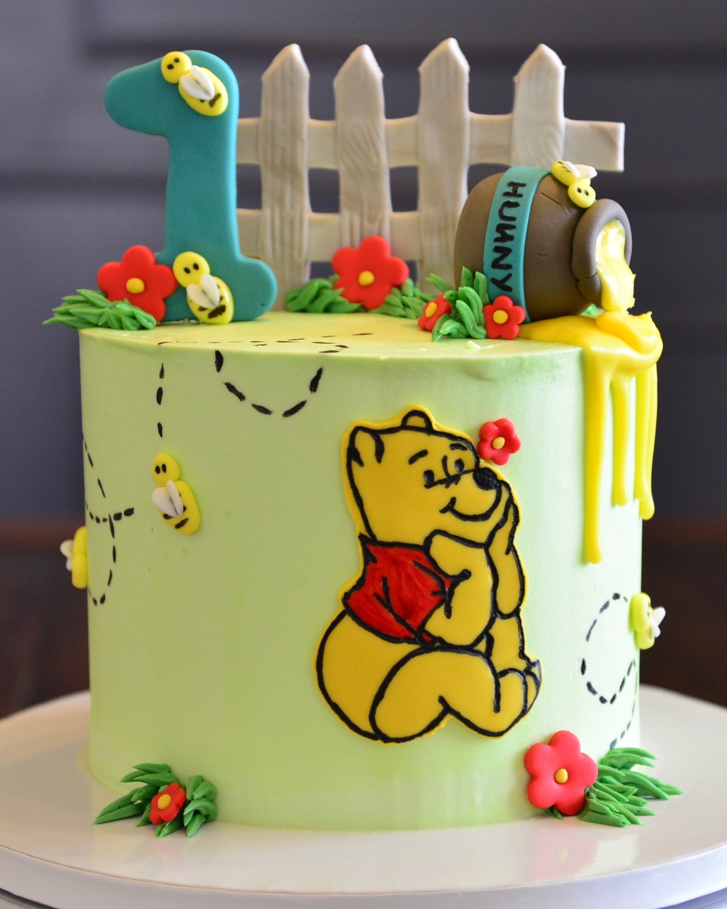 Cake with winnie the pooh theme. Winnie the pooh sitting in the front, an overflowing jar of honey on top and bees and fences.