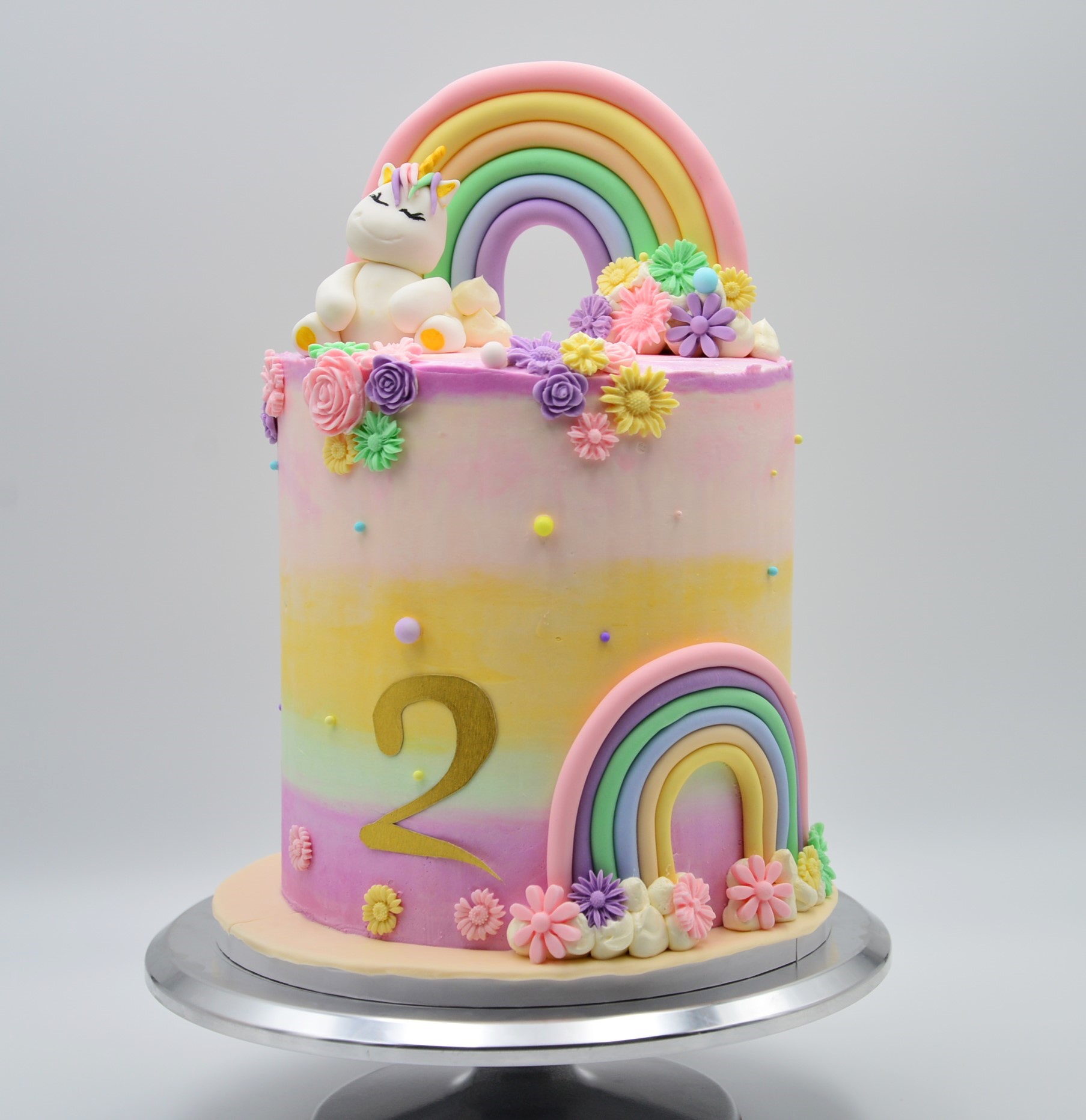 A pastel color rainbow theme tall unicorn cake decorated with flowers, unicorn and rainbows.